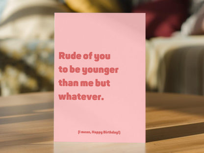 A pink birthday card with the text 'Rude of you to be younger than me but whatever.' and '(I mean, Happy Birthday!)' in a darker pink font.