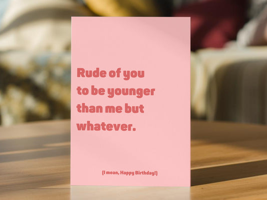A pink birthday card with the text 'Rude of you to be younger than me but whatever.' and '(I mean, Happy Birthday!)' in a darker pink font.