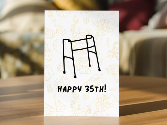 A 35th birthday card with a black line drawing of a zimmerframe and the text 'HAPPY 35TH!'. The card has a subtle gold outline pattern in the background