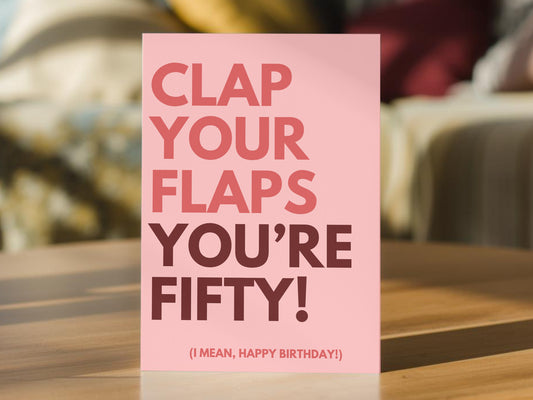 A pink greeting card with bold text reading 'CLAP YOUR FLAPS YOU'RE FIFTY! (I MEAN, HAPPY BIRTHDAY!)' For 50th birthday