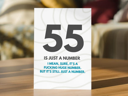 A 55th birthday card standing on a wooden table, featuring a white background with gray wavy line patterns, the large number 55 in dark gray, and the text '55 IS JUST A NUMBER. I MEAN SURE, IT'S A FUCKING HUGE NUMBER, BIT IT'S STILL JUST A NUMBER."