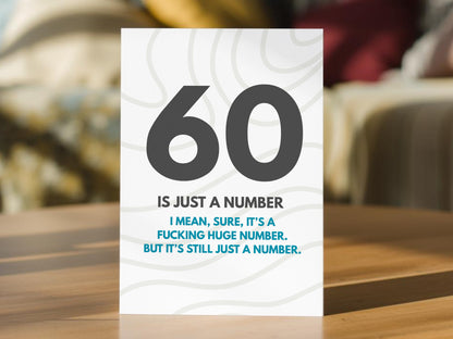 A white 60th birthday card with bold black and blue text reading '60 IS JUST A NUMBER I MEAN, SURE, IT'S A FUCKING HUGE NUMBER. BUT IT'S STILL JUST A NUMBER.