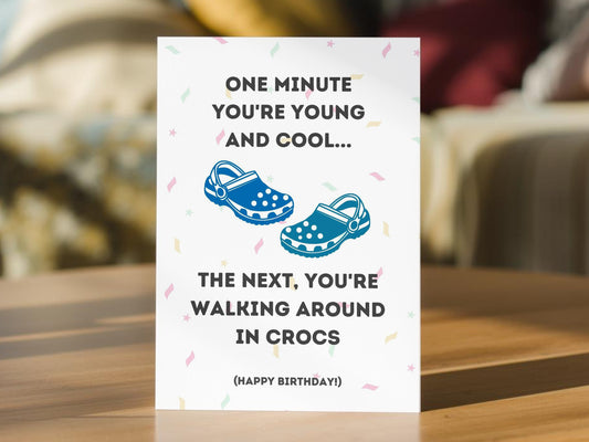 A white birthday card with black text and blue crocs-style shoes. The card reads, 'ONE MINUTE YOU'RE YOUNG AND COOL. THE NEXT, YOU'RE WALKING AROUND IN CROCS (HAPPY BIRTHDAY!)