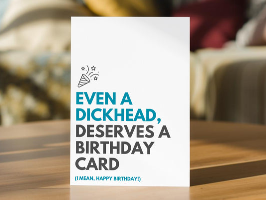 A funny birthday card with bold text reading 'EVEN A DICKHEAD, DESERVES A BIRTHDAY CARD (I MEAN, HAPPY BIRTHDAY!)' in blue and dark gray, standing on a wooden surface with a blurred indoor background.