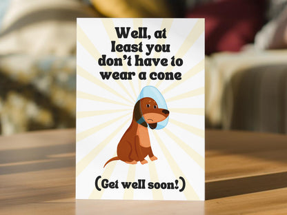 A get well soon greeting card. The card has a white background with yellow sunburst rays, black text reading 'Well, at least you don't have to wear a cone' and '(Get well soon!)', and a cartoon brown dog wearing a light blue cone.