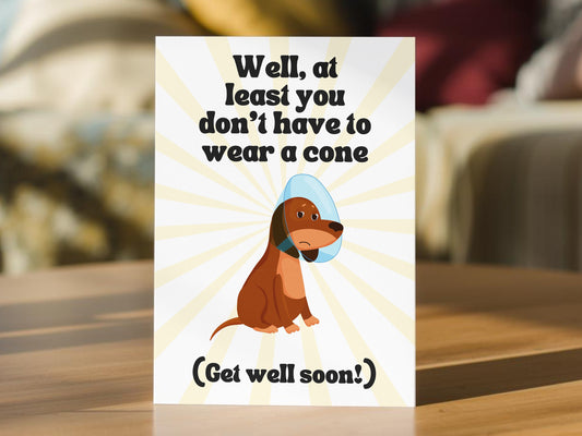 A get well soon greeting card. The card has a white background with yellow sunburst rays, black text reading 'Well, at least you don't have to wear a cone' and '(Get well soon!)', and a cartoon brown dog wearing a light blue cone.
