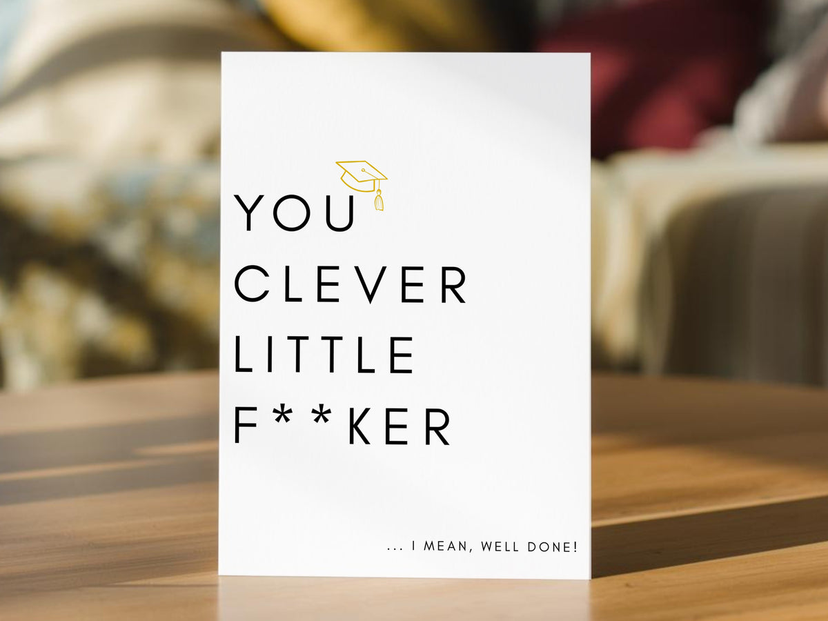A congratulations card with black text reading 'YOU CLEVER LITTLE F**KER' and a small yellow graduation cap icon, standing on a wooden table with a blurred living room background.