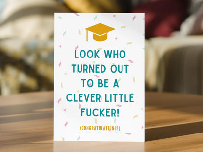 A funny graduation congratulations card. The card features a yellow graduation cap icon, colorful confetti, and teal text that reads 'LOOK WHO TURNED OUT TO BE A CLEVER LITTLE FUCKER! (CONGRATULATIONS!)'.