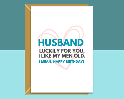 A birthday card for a husband with a kraft envelope, featuring a white card with blue and black text and a light pink heart outline, set against a split teal and light blue background.