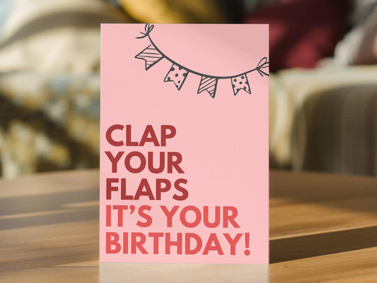 A pink birthday card with the text 'CLAP YOUR FLAPS IT'S YOUR BIRTHDAY!' in red and pink lettering, topped with a black bunting banner illustration