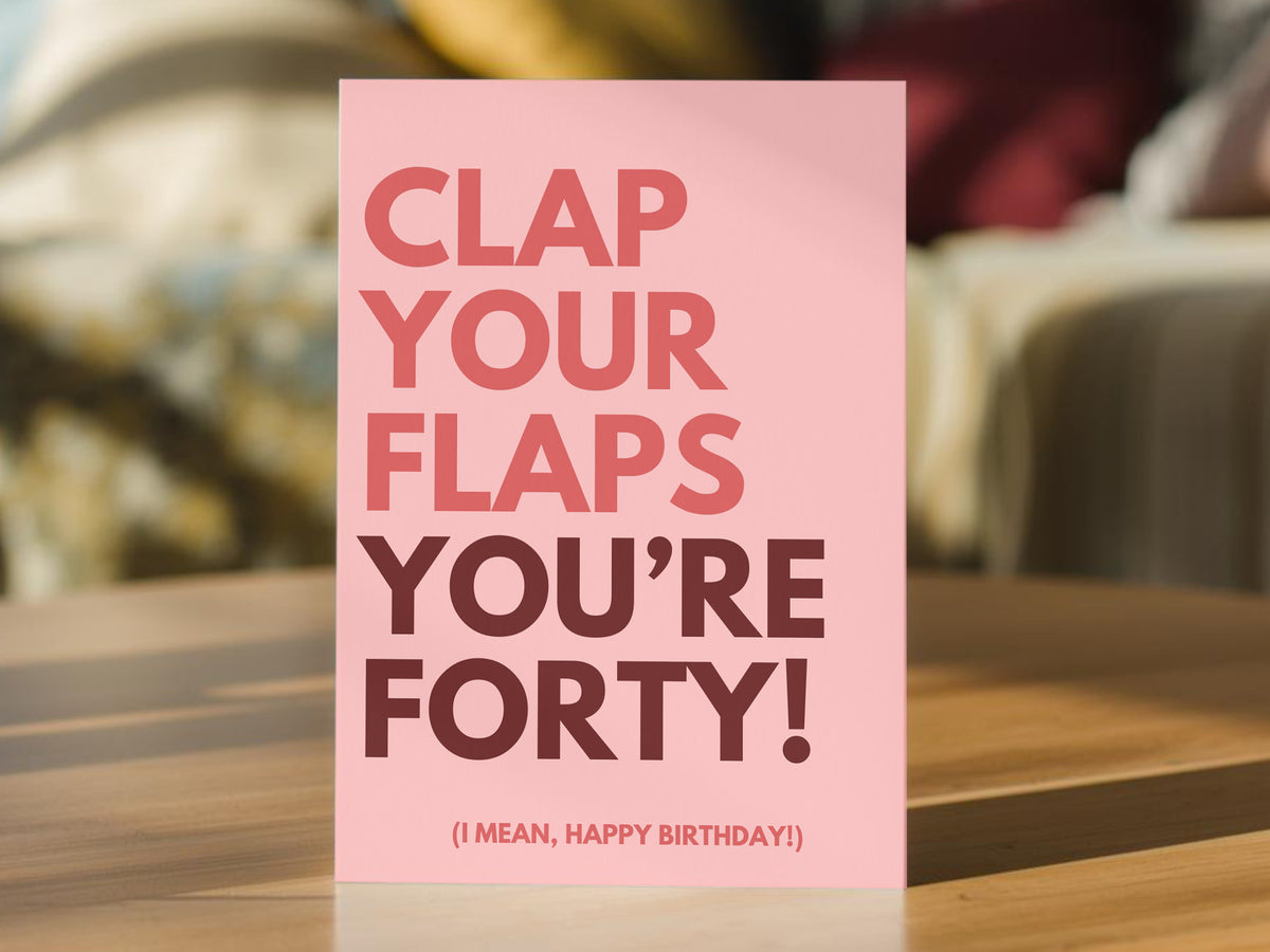 A funny pink greeting card standing upright on a wooden table, with bold text reading 'CLAP YOUR FLAPS YOU'RE FORTY! (I MEAN, HAPPY BIRTHDAY!)' 