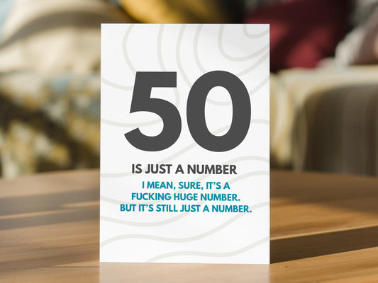 A 50th birthday card standing on a wooden table, featuring a humorous 50th birthday message. The card has a white background with light gray wavy line patterns, a large dark gray '50' at the top, and black and blue text below reading 'IS JUST A NUMBER / I MEAN, SURE, IT'S A FUCKING HUGE NUMBER.