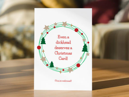 A white Christmas greeting card standing upright on a wooden table, featuring a green wreath with red berries, beige stars, and green pine trees, with red text reading 'Even a dickhead deserves a Christmas Card!' and '(You're welcome!)' at the bottom. 