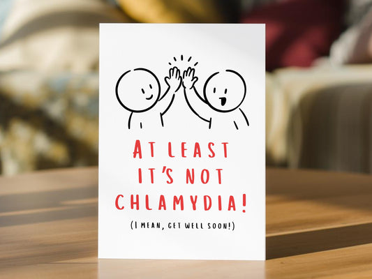 A white get well soon greeting card standing upright on a wooden table, featuring a simple black line drawing of two people high-fiving with the text 'AT LEAST IT'S NOT CHLAMYDIA! (I MEAN, GET WELL SOON!)' in red and black lettering.
